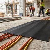 Schwarze geriffelte Gummimatte deckt Stromkabel auf dem Boden einer Baustelle ab, während Handwerker im Hintergrund arbeiten.