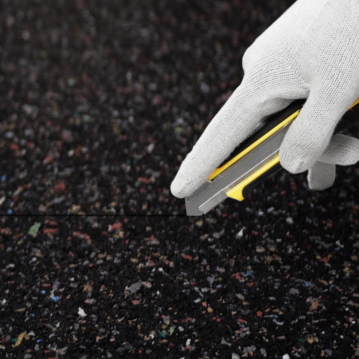 Eine behandschuhte Hand schneidet die schwarze Gummigranulatmatte mit einem Cuttermesser passgenau zu.